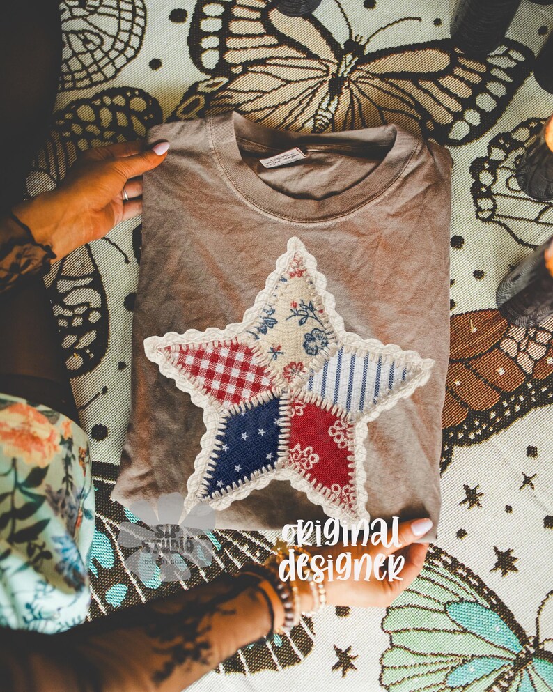 May include: A brown t-shirt with a large, patchwork star applique. The star features red, white, and blue patterned fabrics, including gingham and stripes. The text "original designer" is visible. The shirt is folded on a patterned surface.