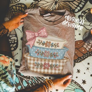May include: A tan-colored t-shirt featuring a stacked design of embroidered casserole dishes. The top dish has a pink polka-dot bow. The text "original designer" is visible on the shirt.