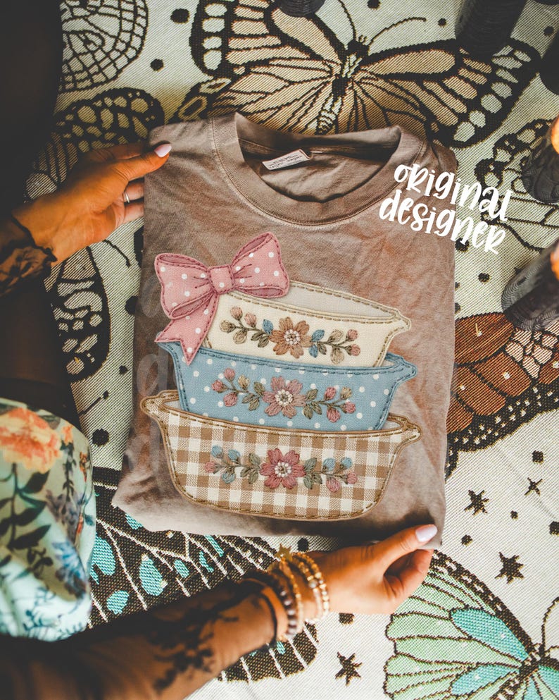 May include: A tan-colored t-shirt featuring a stacked design of embroidered casserole dishes. The top dish has a pink polka-dot bow. The text "original designer" is visible on the shirt.