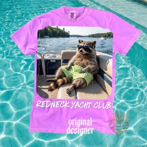 May include: A lilac-colored t-shirt featuring a raccoon wearing sunglasses and green and white striped shorts, lounging on a boat. The shirt has the text "REDNECK YACHT CLUB" and "original designer". A small logo is in the bottom right corner.