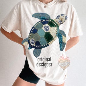 May include: Cream-colored t-shirt featuring a patchwork turtle design in shades of blue and green, embellished with pearls. The shirt has the text "original designer" and a logo with the text "Electric Edge Designs".