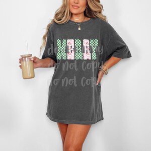 May include: A charcoal gray oversized t-shirt with the word "MERRY" in a block letter design. The letters are a combination of green checkered and pink floral patterns. A clear glass tumbler with a straw is held in the left hand.