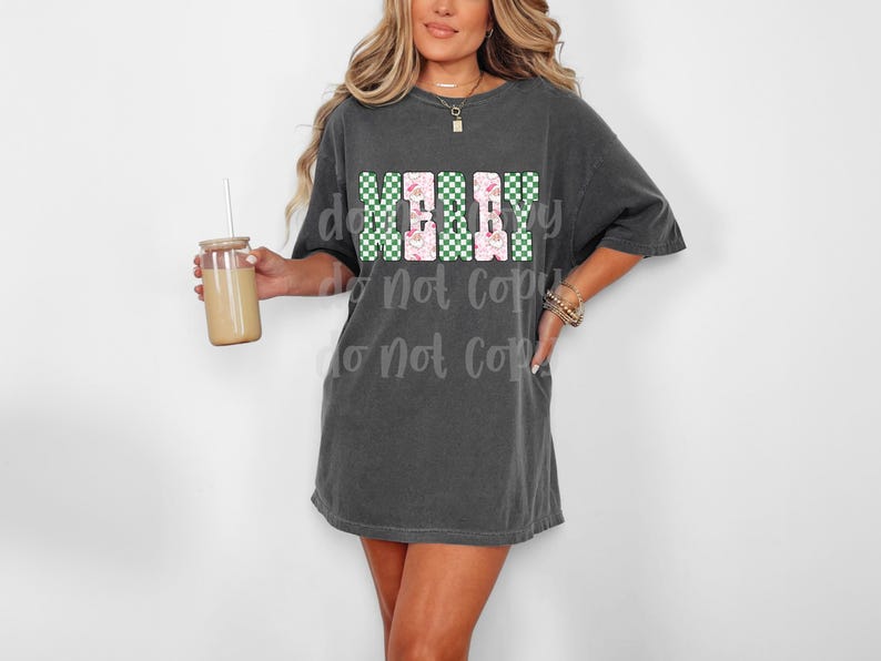 May include: A charcoal gray oversized t-shirt with the word "MERRY" in a block letter design. The letters are a combination of green checkered and pink floral patterns. A clear glass tumbler with a straw is held in the left hand.