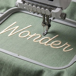 May include: An embroidery machine stitching the word "Wonder" in cream-colored thread onto a sage green fabric. The machine's needle is positioned over the fabric, creating the cursive lettering within a gray frame.