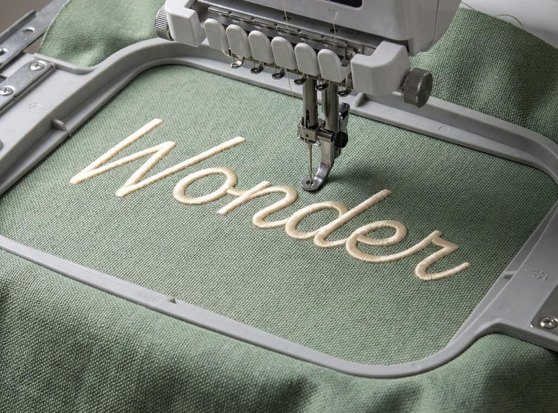 May include: An embroidery machine stitching the word "Wonder" in cream-colored thread onto a sage green fabric. The machine's needle is positioned over the fabric, creating the cursive lettering within a gray frame.
