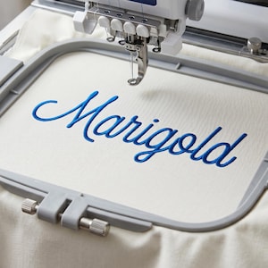May include: An embroidery machine hoop holding a piece of off-white fabric with the word Marigold stitched in a blue cursive font. The machine needle is positioned directly above the center of the fabric within the gray plastic frame.