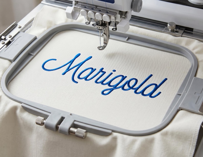 May include: An embroidery machine hoop holding a piece of off-white fabric with the word Marigold stitched in a blue cursive font. The machine needle is positioned directly above the center of the fabric within the gray plastic frame.