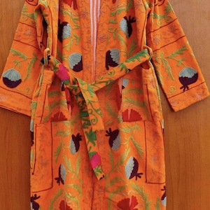 May include: A bright orange long-sleeved robe featuring an all-over embroidered floral pattern in shades of red, purple, and green. The garment includes a matching tie belt at the waist, two front patch pockets, and a white and orange striped lining.