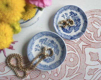 May include: Two small blue and white decorative plates with gold jewelry. One plate holds gold hoop earrings, the other holds a gold chain necklace and a gold clasp. The plates have a classic blue and white design.