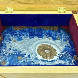 Puede incluir: Caja de madera con tapa con bisagras, herrajes de latón e interior forrado de terciopelo. Contiene una obra de arte en resina con tonos azules, blancos y dorados, que se asemeja a un paisaje abstracto. Engranajes decorativos en las esquinas.
