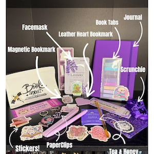 Puede incluir: Una colección de accesorios con temática de libros, que incluye un diario morado, pestañas de libros, pegatinas, clips y una scrunchie. Otros artículos incluyen una mascarilla, un marcapáginas de corazón de cuero y un marcapáginas magnético. Sobre fondo negro.
