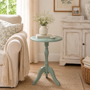 May include: A round, light blue side table with a three-legged base. The table is topped with a white vase of flowers, a book, and a small white bowl. The table is approximately 24 inches tall.