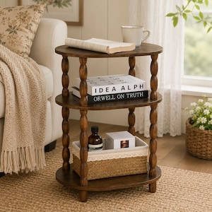 May include: A three-tiered, dark brown wooden side table with a round top and shelves. The table features turned legs and holds books, a mug, and a woven basket. The books read "IDEA ALPH" and "ORWELL ON TRUTH".
