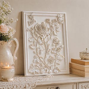 May include: A framed, white relief sculpture of roses. The artwork features detailed, three-dimensional roses, leaves, and stems. The frame is also white, and the piece is displayed on a white surface with other decorative items.