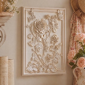 May include: A rectangular, off-white framed wall art piece featuring a raised relief of a rose and foliage in a gold color. The artwork has a vintage, romantic aesthetic, with detailed leaves and petals.