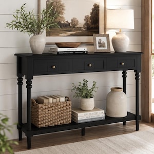 May include: A black console table with three drawers and gold knobs. The table has a lower shelf and turned legs. It is decorated with a vase, books, a lamp, and a framed picture. A woven basket and potted plants are also on display.
