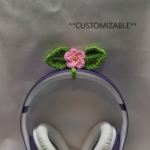May include: A crocheted headphone accessory with a pink flower and green leaves, attached to purple and white headphones. The text "**CUSTOMIZABLE**" is visible. The headphones are a popular audio accessory.