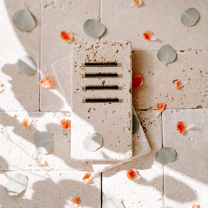 Op de afbeelding: Vier rijen zwarte wimperextensies worden weergegeven op een beige stenen tegel. De tegel is omringd door kleine, lichtroze bloemblaadjes en groene bladeren.