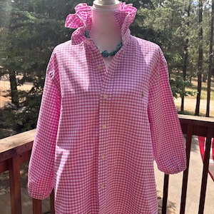 May include: A long-sleeved button-down shirt featuring a pink and white gingham check pattern. The shirt has a high, ruffled collar and a small embroidered logo on the left chest. It is displayed on a mannequin wearing a turquoise beaded necklace.