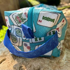 May include: A square, zippered bag with a blue handle and a Mahjong tile pattern. The bag's fabric is light blue with white tiles featuring red, green, and black symbols. The zipper and handle are a darker shade of blue.