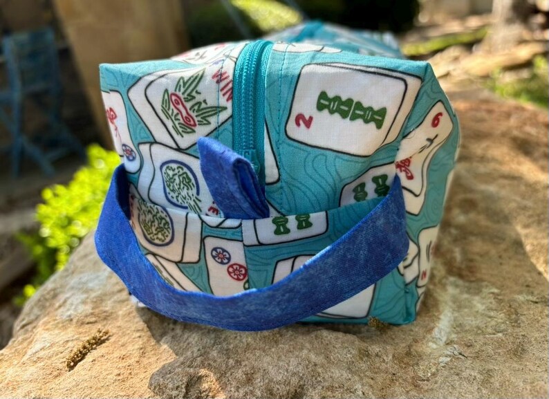 May include: A square, zippered bag with a blue handle and a Mahjong tile pattern. The bag's fabric is light blue with white tiles featuring red, green, and black symbols. The zipper and handle are a darker shade of blue.