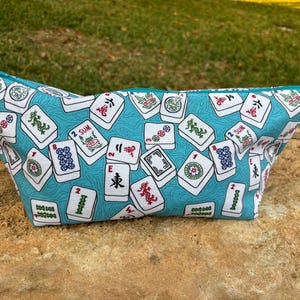 May include: A turquoise zippered pouch featuring a pattern of white Mahjong tiles with red, green, and black symbols. The pouch is made of fabric and has a teal zipper and trim. The bag is sitting on a rock.