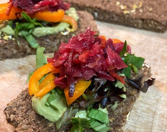 May include: A close-up of a sandwich with a base of dark bread topped with avocado spread, red cabbage, orange bell peppers, and fresh greens.