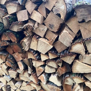 May include: A close-up of a stack of firewood. The wood is a mix of light and dark brown colors, with some pieces showing the bark still attached. The wood is neatly stacked, with the pieces arranged in a pattern.