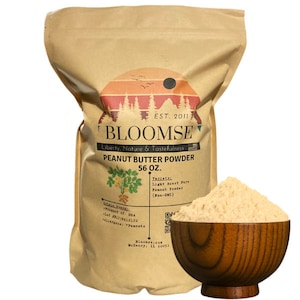 May include: A brown paper bag with a white label that reads "BLOOMSE Liberty, Nature & Tastefulness PEANUT BUTTER POWDER 56 OZ. Variety: Light Roast Pure Peanut Powder (Non-GMO)". A wooden bowl filled with peanut butter powder sits in front of the bag.