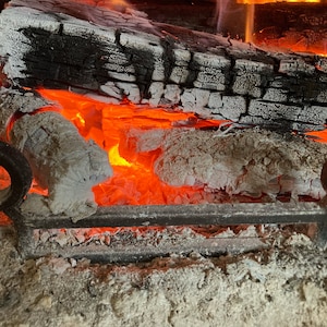 May include: Close-up of a fireplace with burning logs and embers. The logs are partially burned and have a white ash coating. The embers are glowing red and orange.