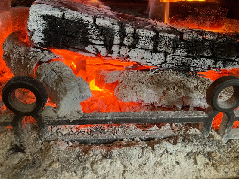 May include: Close-up of a fireplace with burning logs and embers. The logs are partially burned and have a white ash coating. The embers are glowing red and orange.