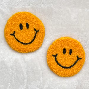 May include: Two round, yellow coasters with a smiley face design. Each coaster features a textured, fuzzy surface with a black, smiling mouth and two black eyes. The coasters are placed on a white, textured surface.