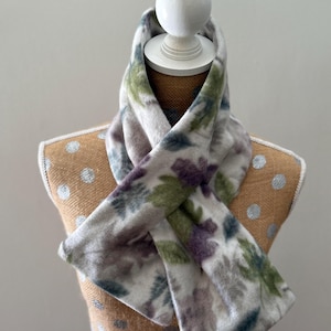 May include: A patterned scarf on a mannequin. The scarf has a white background with a floral pattern in green, purple, and grey. The scarf is draped around the mannequin's neck, highlighting its design and texture. The mannequin is on a brown surface.