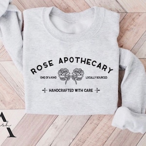K&ouml;nnte beinhalten: Grauer Pullover mit dem Text "ROSE APOTHECARY" in schwarzer Schrift. Der Text ist von zwei stilisierten Rosen und den Worten "ONE OF A KIND" und "LOCALLY SOURCED" umgeben. Der Text "HANDCRAFTED WITH CARE" befindet sich unter den Rosen.