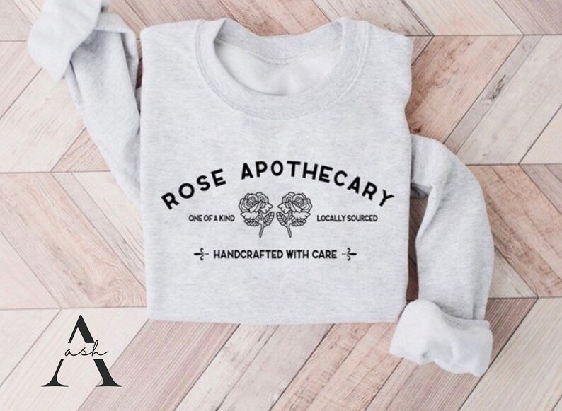K&ouml;nnte beinhalten: Grauer Pullover mit dem Text "ROSE APOTHECARY" in schwarzer Schrift. Der Text ist von zwei stilisierten Rosen und den Worten "ONE OF A KIND" und "LOCALLY SOURCED" umgeben. Der Text "HANDCRAFTED WITH CARE" befindet sich unter den Rosen.