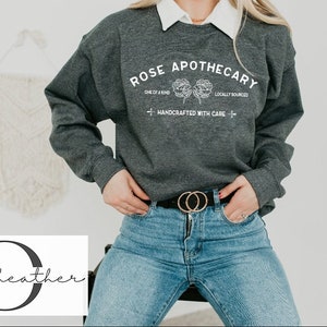 K&ouml;nnte beinhalten: Ein dunkelgrauer Sweatshirt mit dem Text "ROSE APOTHECARY" in Wei&szlig;, mit einem floralen Design und den Worten "ONE OF A KIND", "LOCALLY SOURCED" und "HANDCRAFTED WITH CARE".