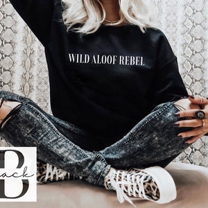 May include: A black sweatshirt with the text "WILD ALOOF REBEL" printed in white on the front. The model is wearing ripped jeans and leopard print sneakers.
