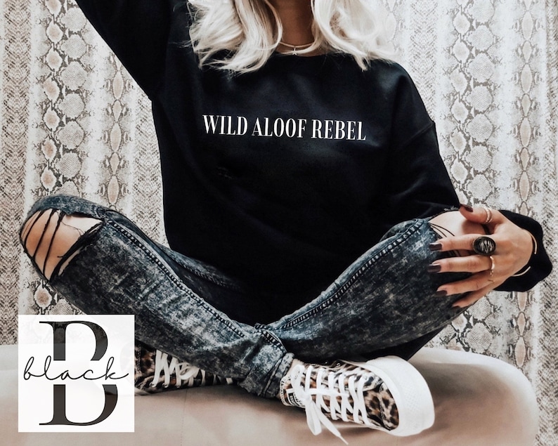 May include: A black sweatshirt with the text "WILD ALOOF REBEL" printed in white on the front. The model is wearing ripped jeans and leopard print sneakers.
