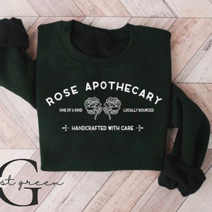K&ouml;nnte beinhalten: Ein dunkelgr&uuml;ner Pullover mit dem Text "ROSE APOTHECARY" in Wei&szlig;, mit zwei stilisierten Rosen und den S&auml;tzen "ONE OF A KIND" und "LOCALLY SOURCED". Der Text "HANDCRAFTED WITH CARE" ist ebenfalls in Wei&szlig; enthalten.