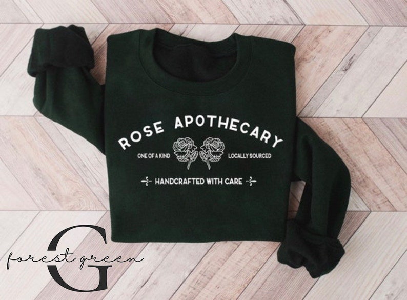 K&ouml;nnte beinhalten: Ein dunkelgr&uuml;ner Pullover mit dem Text "ROSE APOTHECARY" in Wei&szlig;, mit zwei stilisierten Rosen und den S&auml;tzen "ONE OF A KIND" und "LOCALLY SOURCED". Der Text "HANDCRAFTED WITH CARE" ist ebenfalls in Wei&szlig; enthalten.