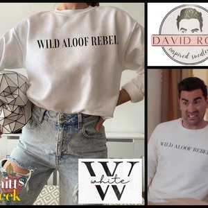 May include: White crewneck sweatshirt with the text "WILD ALOOF REBEL" printed in black on the front. The sweatshirt is shown on a model wearing ripped light blue jeans.