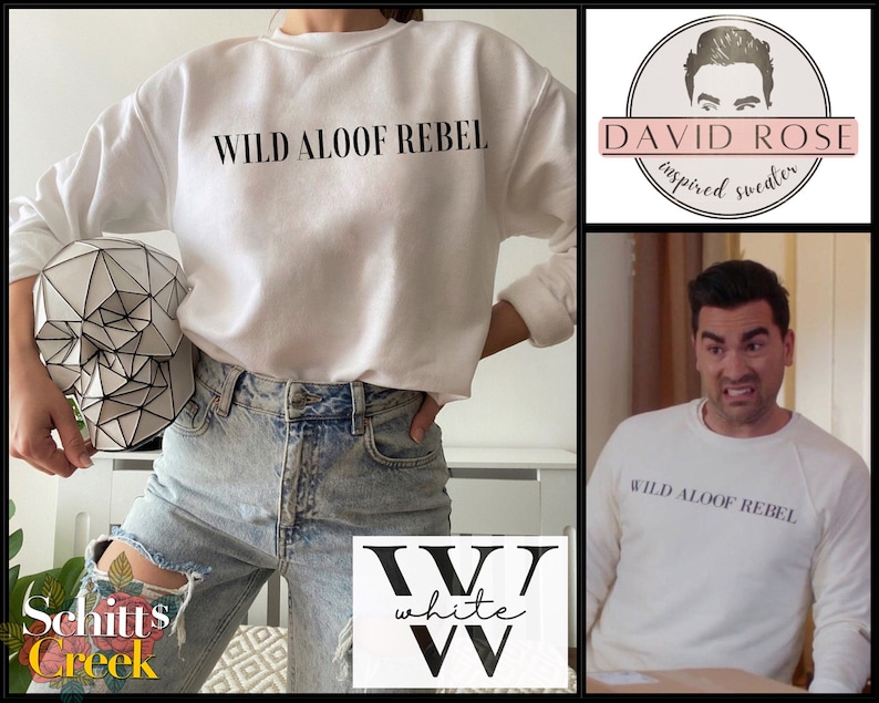 May include: White crewneck sweatshirt with the text "WILD ALOOF REBEL" printed in black on the front. The sweatshirt is shown on a model wearing ripped light blue jeans.