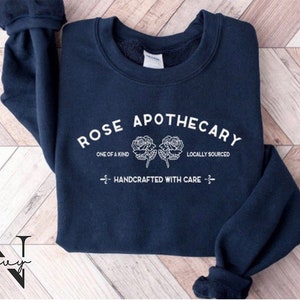 K&ouml;nnte beinhalten: Marineblauer Sweatshirt mit wei&szlig;em Text, der "ROSE APOTHECARY", "ONE OF A KIND", "LOCALLY SOURCED" und "HANDCRAFTED WITH CARE" lautet. Der Text ist in einem Kreisbogen mit zwei stilisierten Rosen in der Mitte.