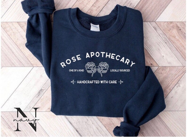 K&ouml;nnte beinhalten: Marineblauer Sweatshirt mit wei&szlig;em Text, der "ROSE APOTHECARY", "ONE OF A KIND", "LOCALLY SOURCED" und "HANDCRAFTED WITH CARE" lautet. Der Text ist in einem Kreisbogen mit zwei stilisierten Rosen in der Mitte.