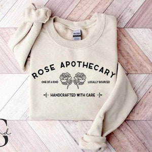 May include: A beige sweatshirt with the text "ROSE APOTHECARY" in black lettering. The text is surrounded by two stylized roses and the words "ONE OF A KIND" and "LOCALLY SOURCED". Below the roses is the text "HANDCRAFTED WITH CARE".