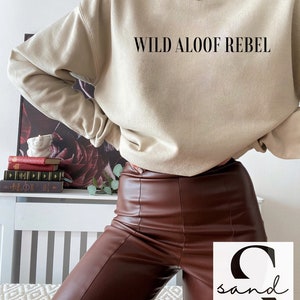 May include: A beige sweatshirt with the text "WILD ALOOF REBEL" printed on the front. The model is wearing brown leather pants.