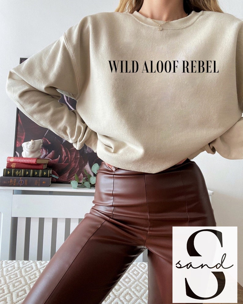 May include: A beige sweatshirt with the text "WILD ALOOF REBEL" printed on the front. The model is wearing brown leather pants.