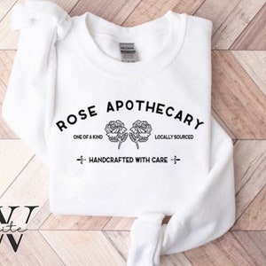 K&ouml;nnte beinhalten: Wei&szlig;er Pullover mit schwarzem Text, der "ROSE APOTHECARY", "ONE OF A KIND", "LOCALLY SOURCED" und "HANDCRAFTED WITH CARE" lautet. Der Text ist von zwei schwarzen Rosen umgeben.