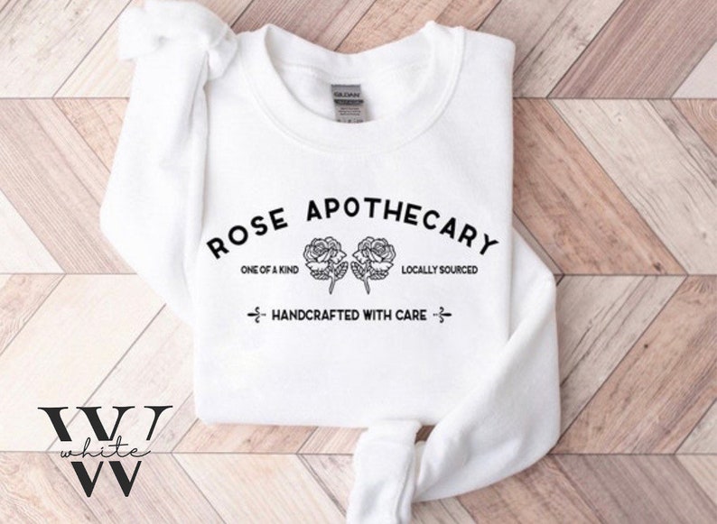 K&ouml;nnte beinhalten: Wei&szlig;er Pullover mit schwarzem Text, der "ROSE APOTHECARY", "ONE OF A KIND", "LOCALLY SOURCED" und "HANDCRAFTED WITH CARE" lautet. Der Text ist von zwei schwarzen Rosen umgeben.