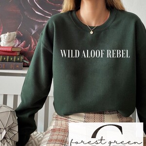 May include: A forest green sweatshirt with the text "WILD ALOOF REBEL" printed on the front. The sweatshirt is worn by a person wearing a plaid skirt.
